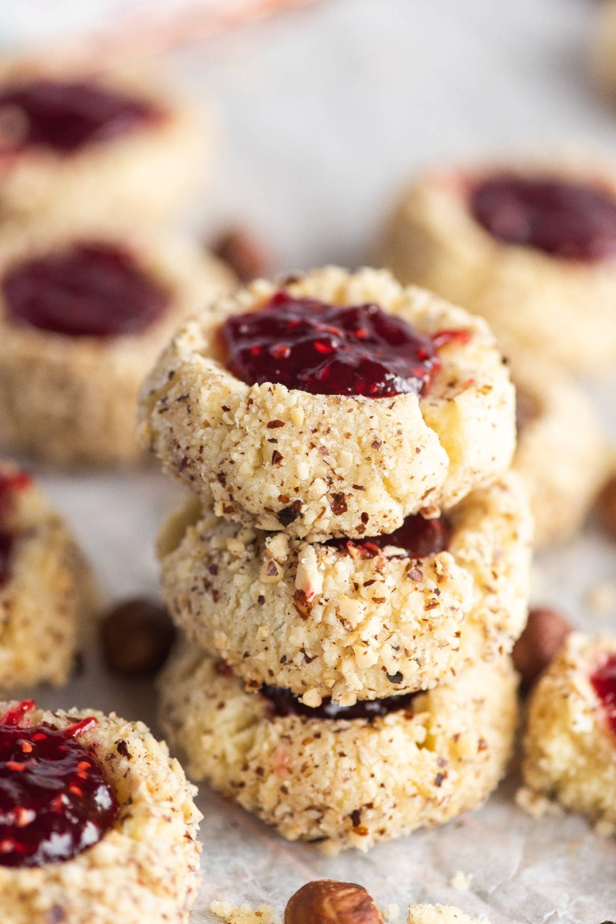 Three classic thumbprint cookies with chopped nuts and red jam stacked, with more cookies and nuts in the background.