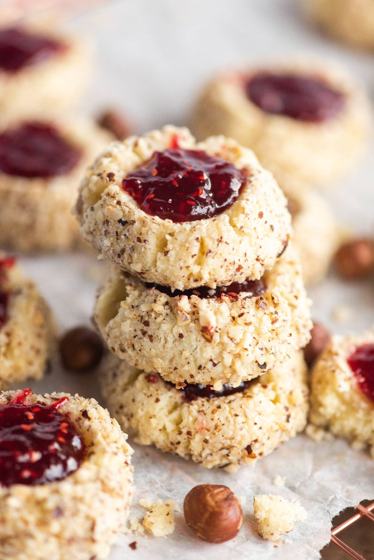 A stack of hazelnut thumbprint cookies with jam sits surrounded by more classic thumbprint cookies and whole hazelnuts, featuring a red jam filling and chopped nuts for added crunch.