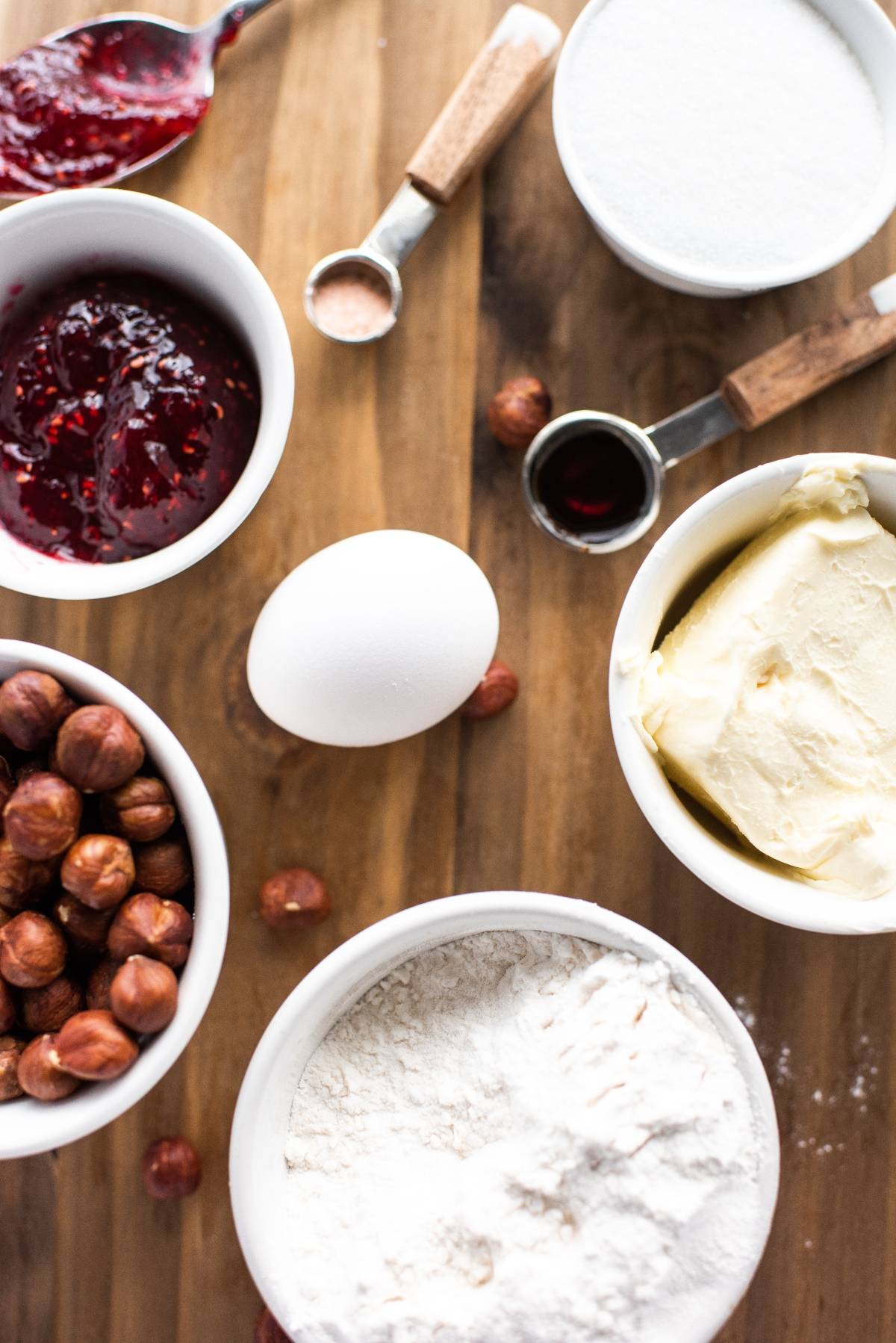 Baking ingredients on a wooden surface: flour, egg, butter, sugar, jam, hazelnuts, vanilla, and salt—all you need to make delicious hazelnut thumbprint cookies with jam.