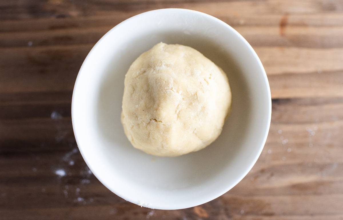 A ball of dough in a white bowl sits on a wooden surface, ready to be shaped into classic thumbprint cookies.