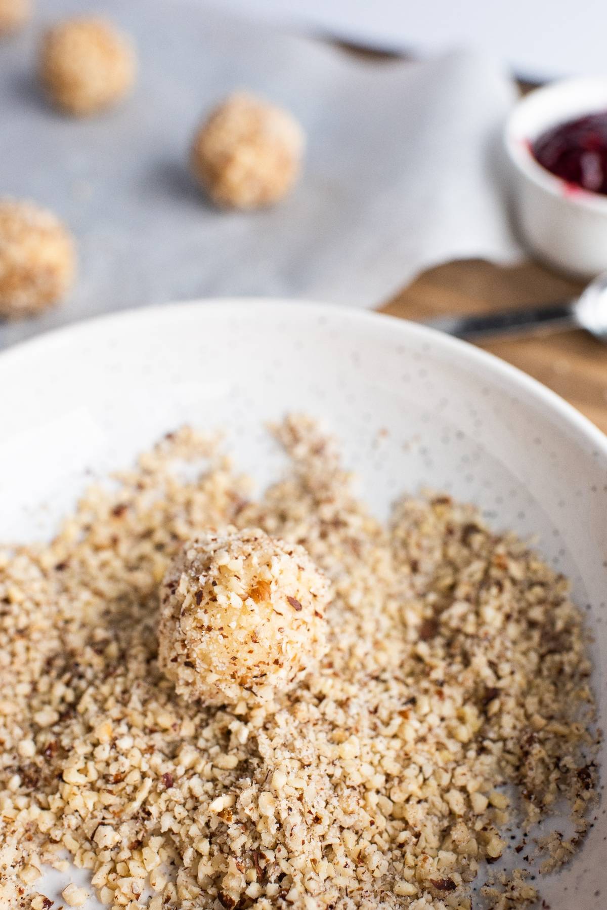 A round ball being coated in chopped nuts on a plate, with more hazelnut thumbprint cookies with jam in the background.