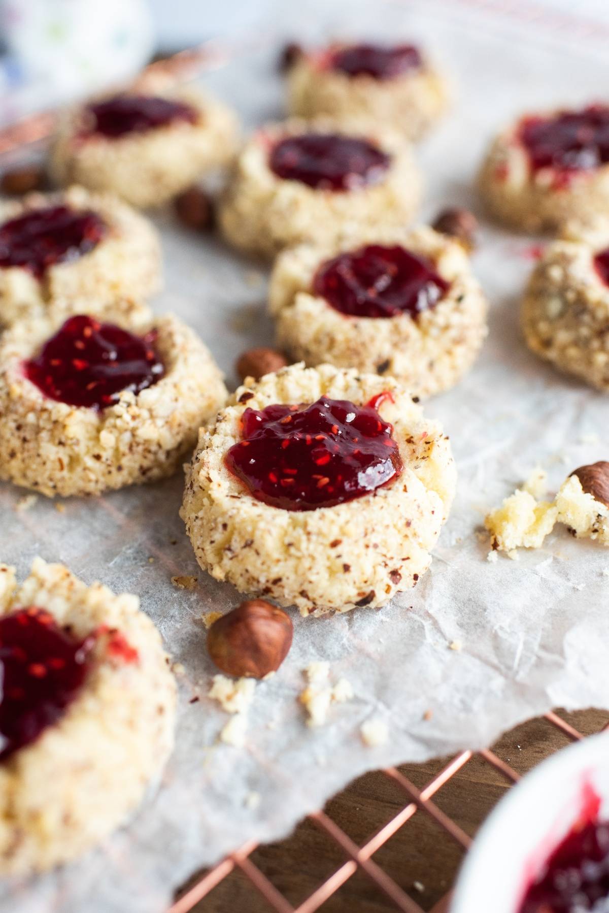 Nut-studded hazelnut thumbprint cookies with jam are filled with red raspberry jam and arranged on parchment paper, surrounded by scattered hazelnuts.