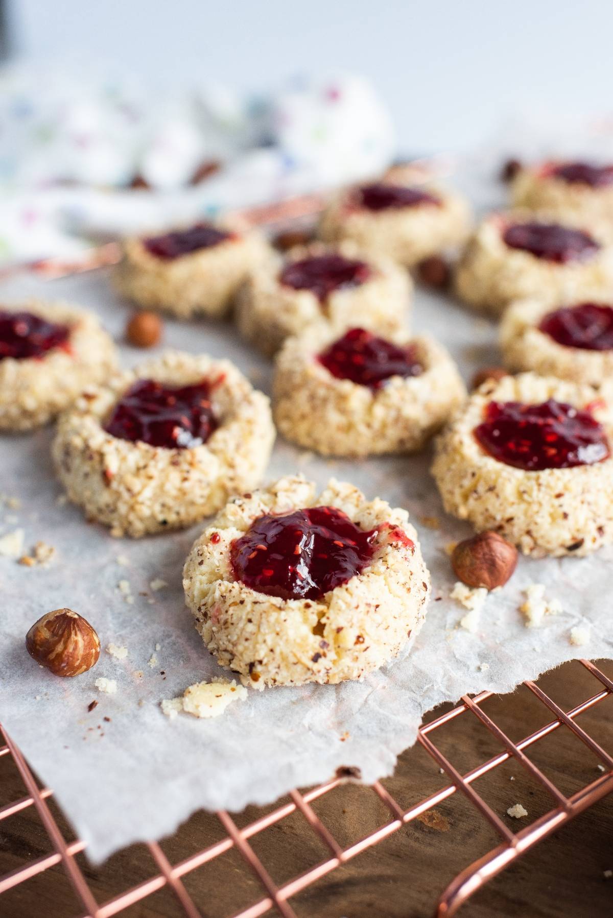 Jam-filled classic thumbprint cookies with a crumbly coating cool on a wire rack, surrounded by scattered hazelnuts.