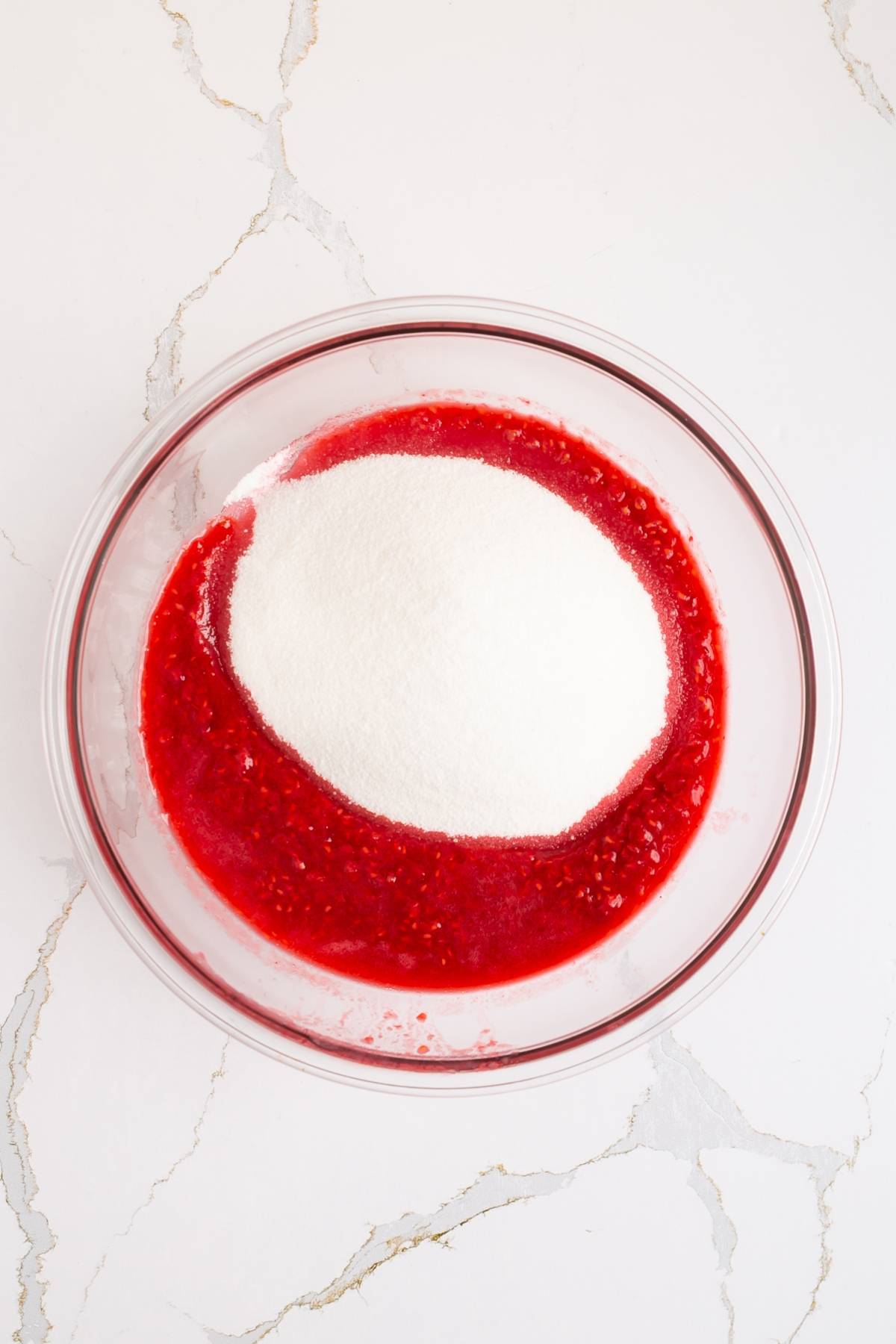 A glass bowl with raspberry puree and a mound of white sugar on a white marble surface.