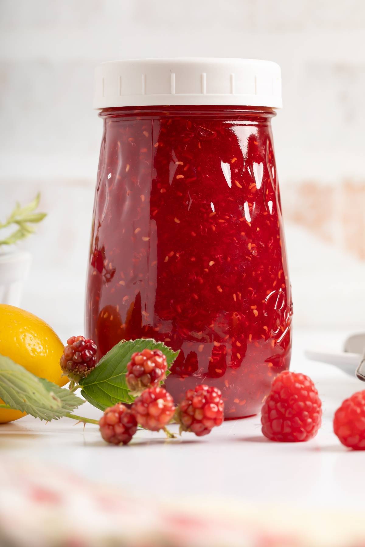 A jar of raspberry jam with fresh raspberries and a lemon on a white surface.