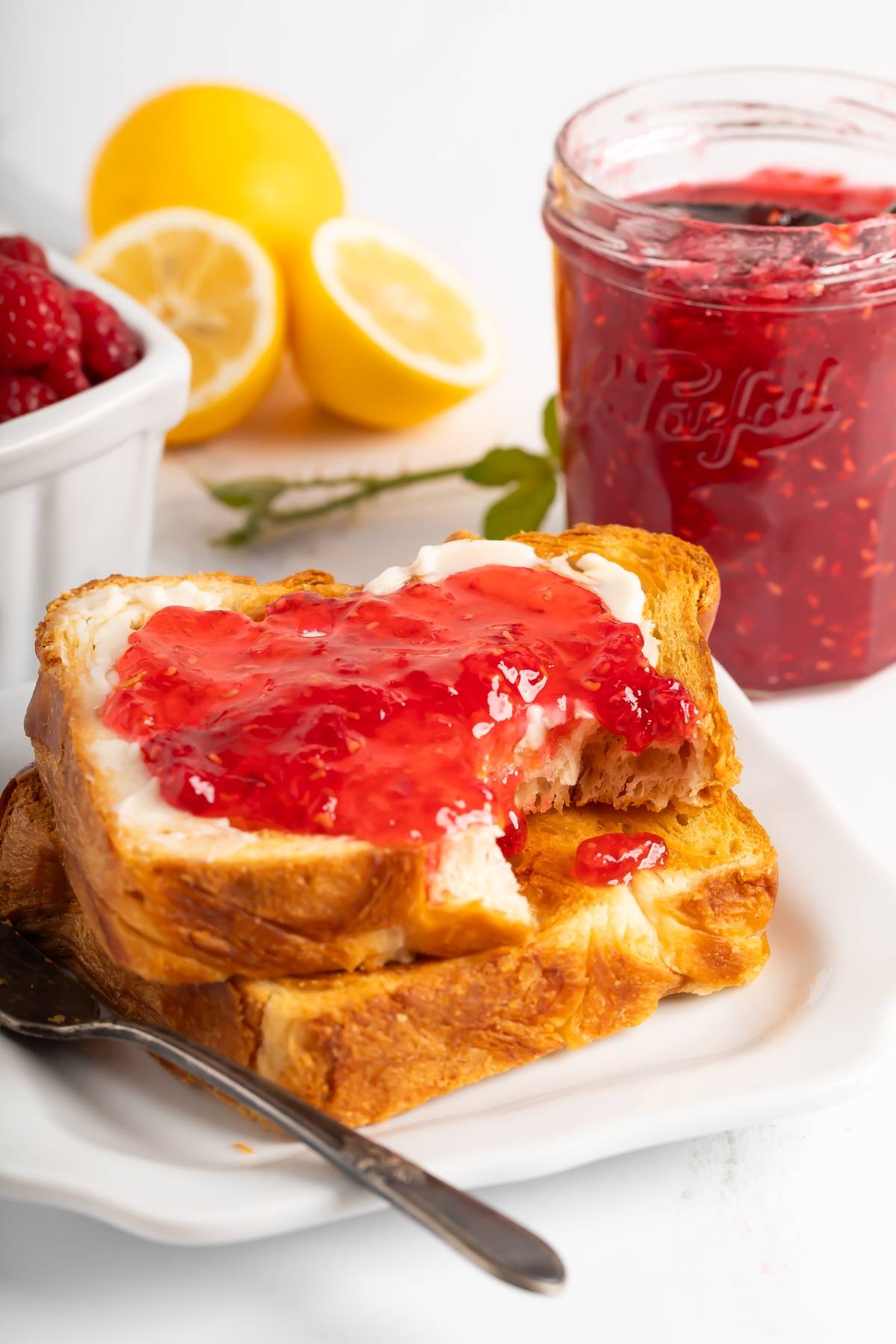 Two slices of toast with butter and raspberry jam, next to a jar of jam, lemons, and fresh raspberries.