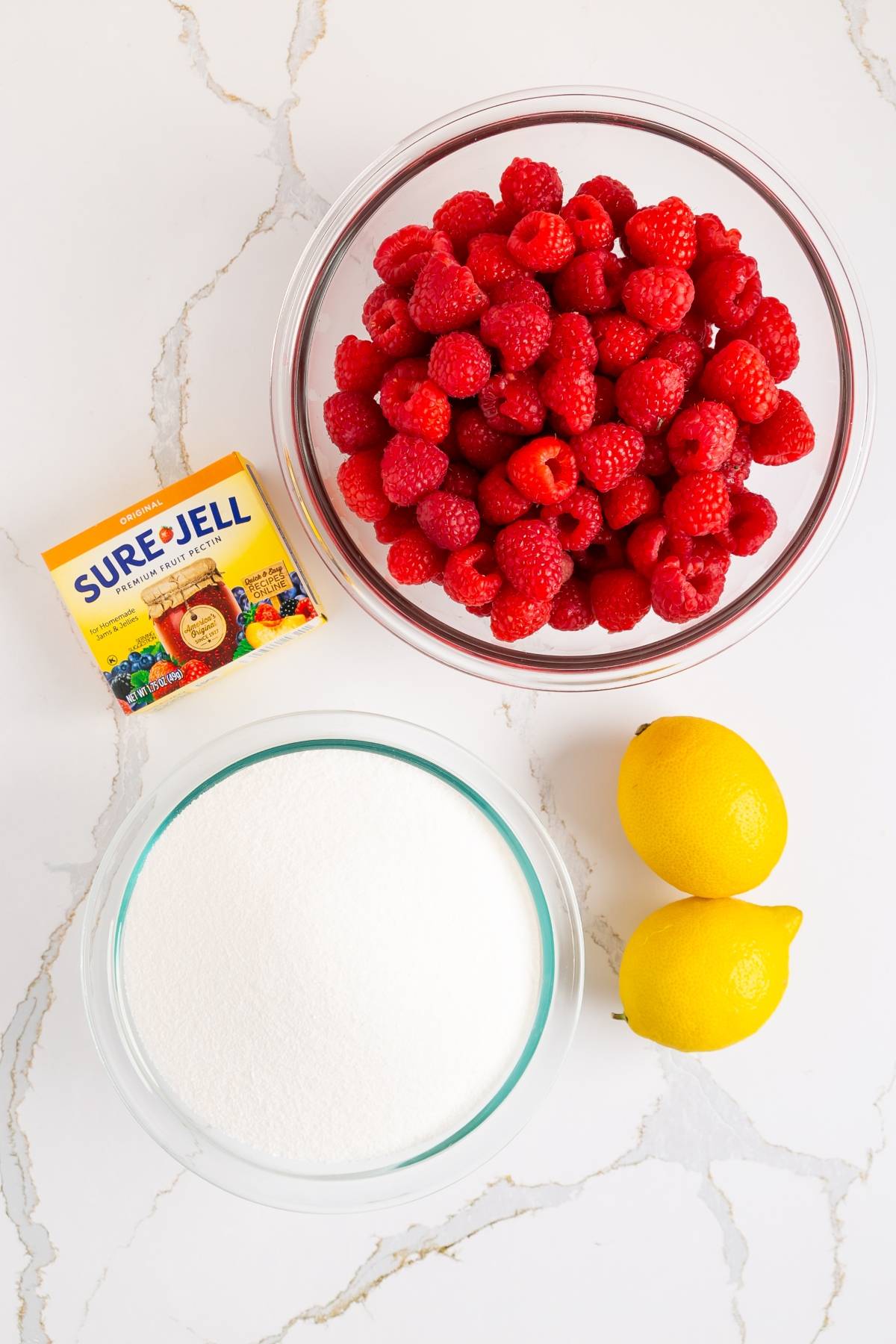 A bowl of raspberries, two lemons, a bowl of sugar, and a box of Sure-Jell on a white countertop.