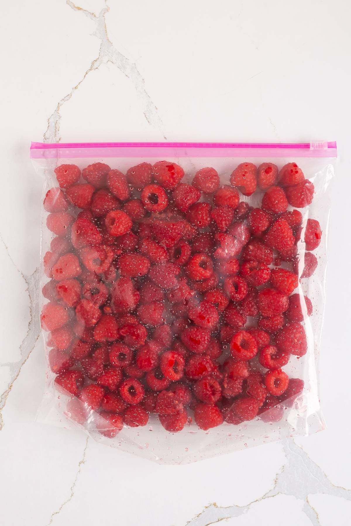 A plastic zipper bag filled with fresh raspberries on a white marble surface.