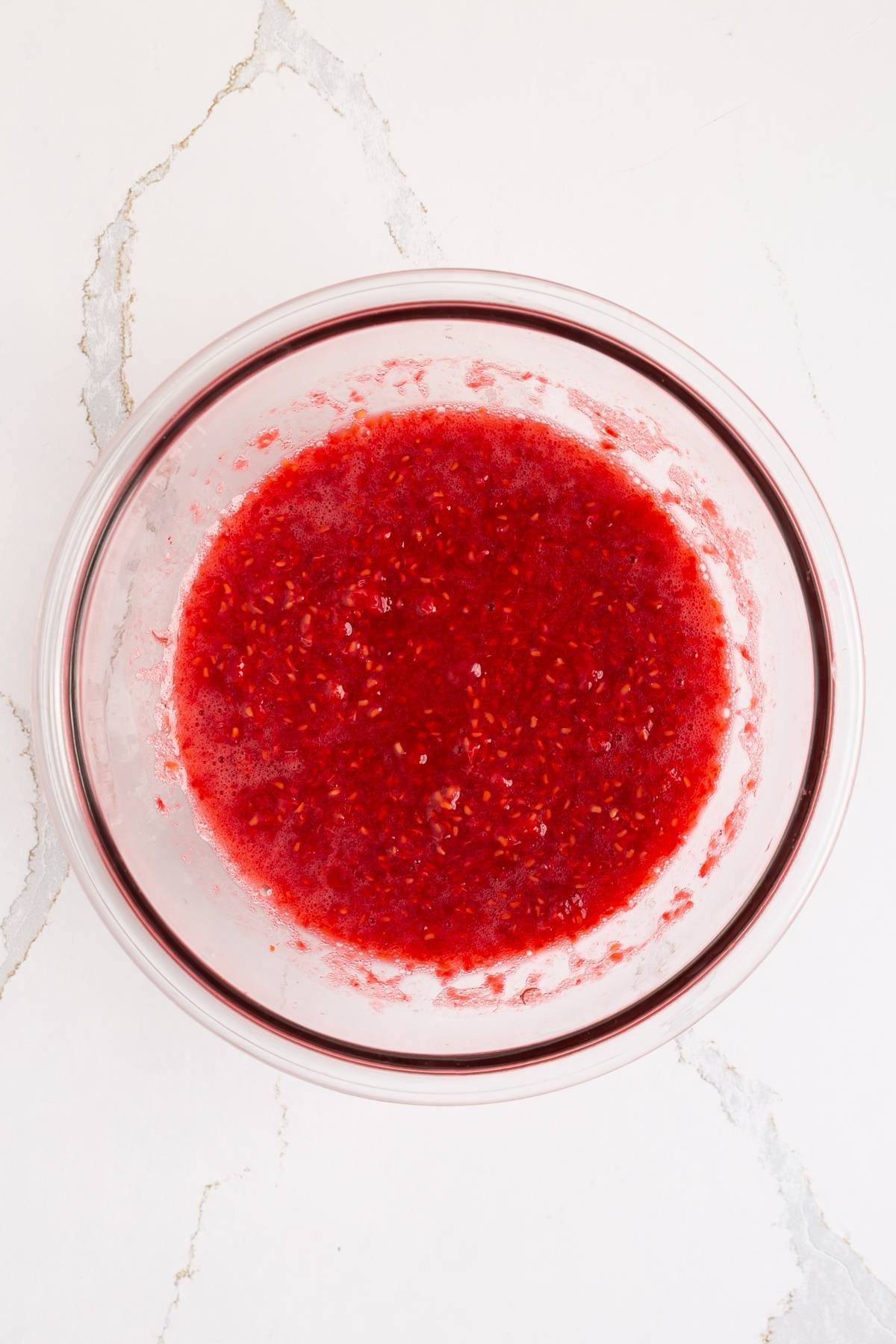 A glass bowl filled with mashed raspberries on a white marble surface.