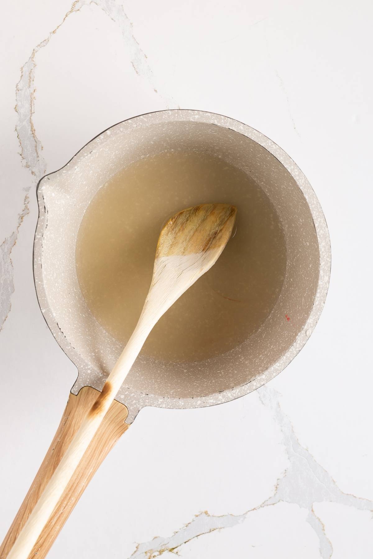 A saucepan with a wooden spoon stirring a clear liquid on a white marble surface.