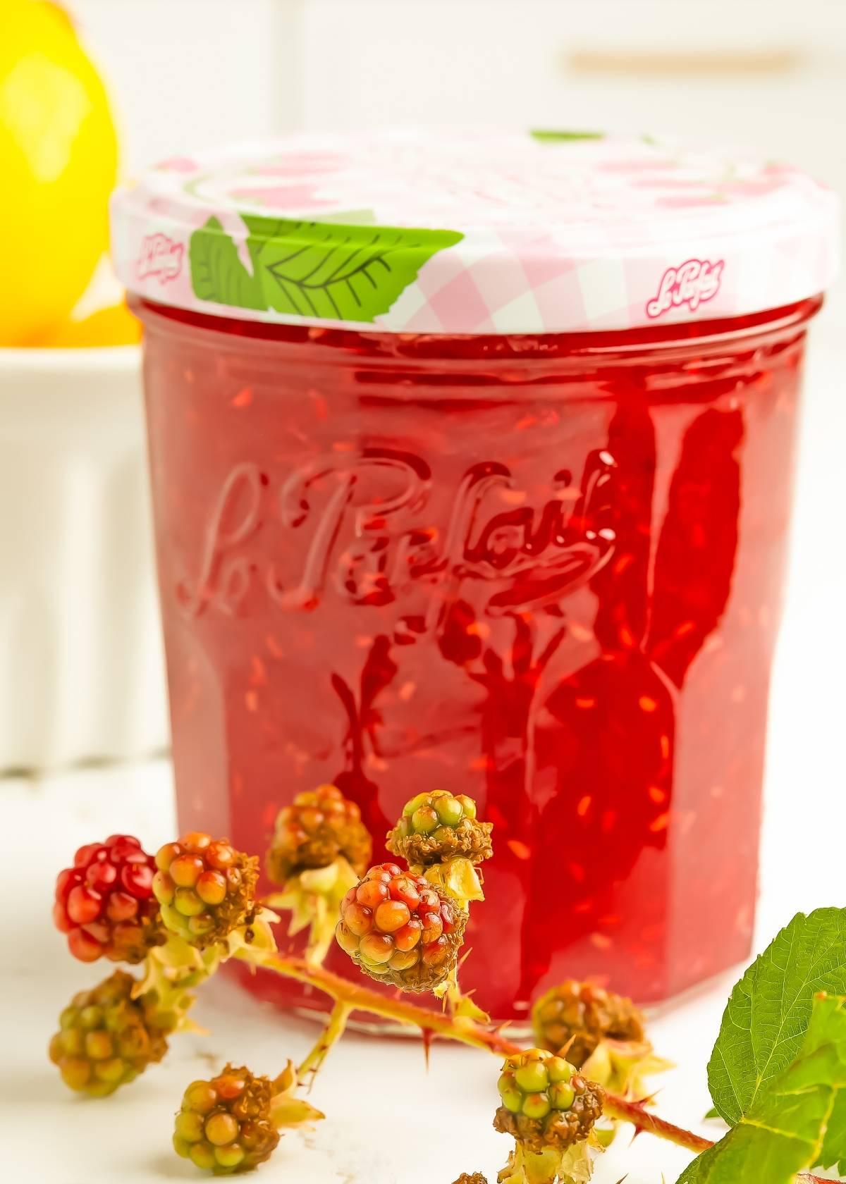 A jar of raspberry jam with a white and pink lid, next to some fresh unripe blackberries.