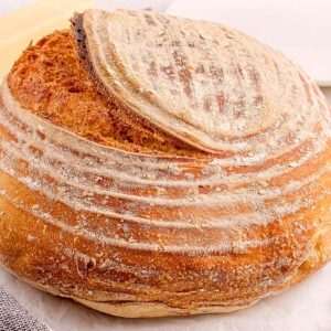 Round loaf of beginner's sourdough bread on a table beside a bread knife, striped towel, butter, and a white plate.