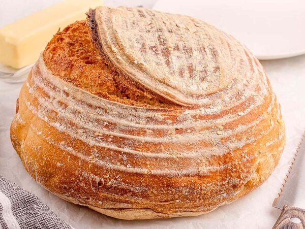 Round loaf of beginner's sourdough bread on a table beside a bread knife, striped towel, butter, and a white plate.
