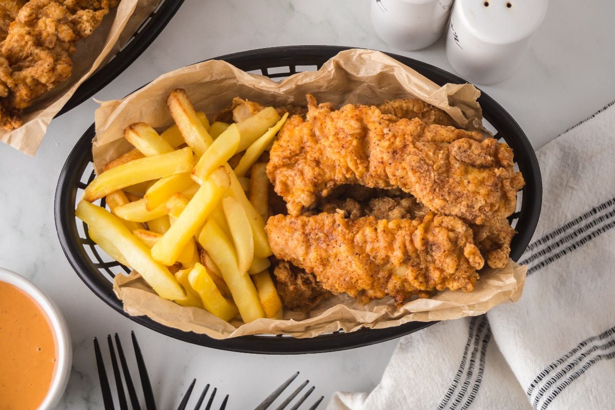 Basket of crispy spicy chicken tenders and golden French fries on parchment paper, with sauce and utensils nearby.