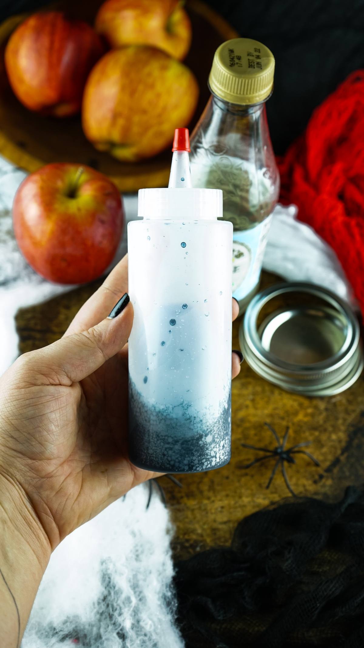 A hand holds a squeeze bottle with dark liquid—perfect for creating Vampire Kiss Halloween Vial Shooter alcoholic shots for Halloween, with apples and a bottle in the background.