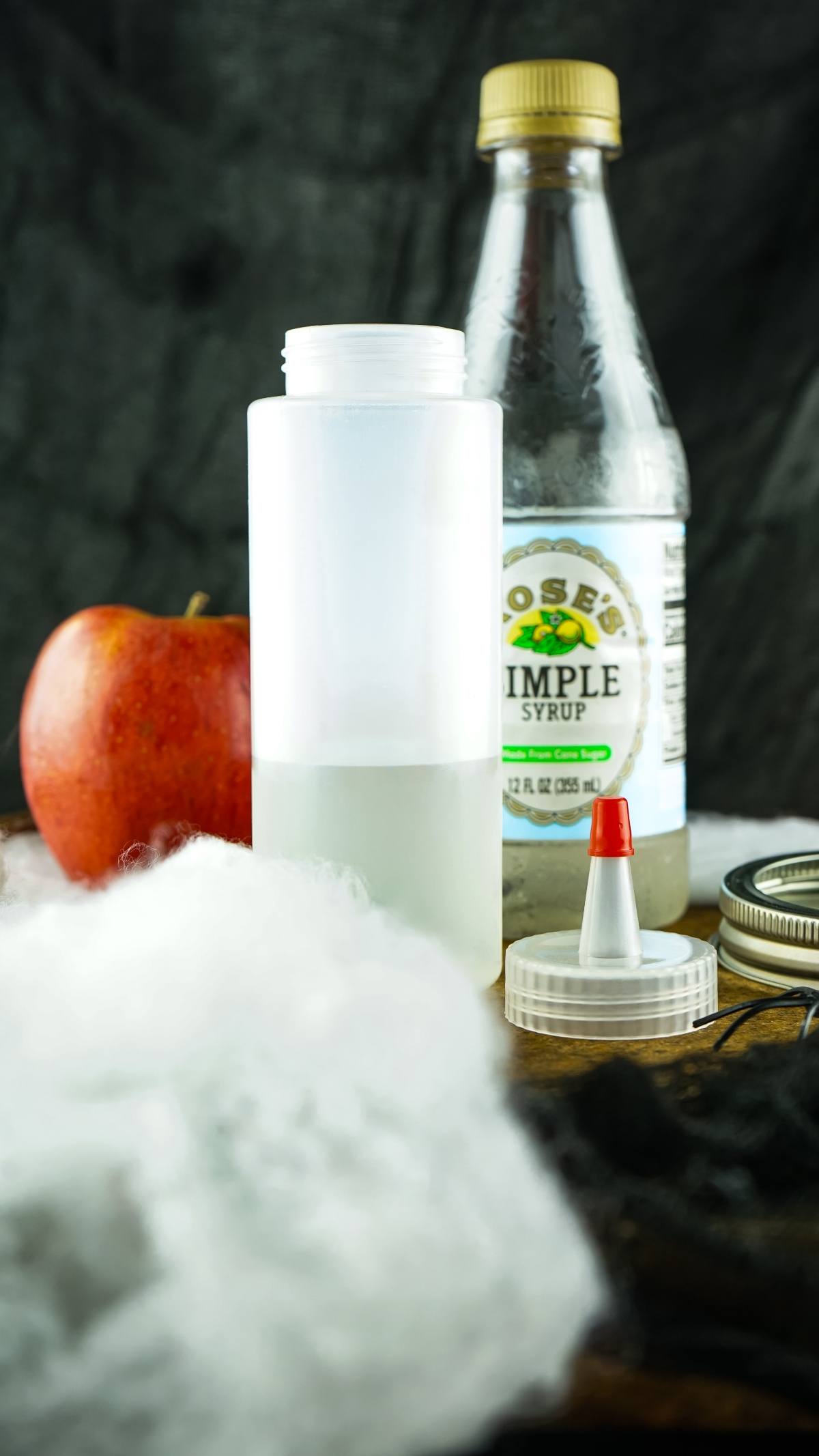 A plastic bottle, apple, simple syrup bottle, cotton, and dropper cap arranged on a table—perfect props for crafting creative alcoholic shots for Halloween at your next party.
