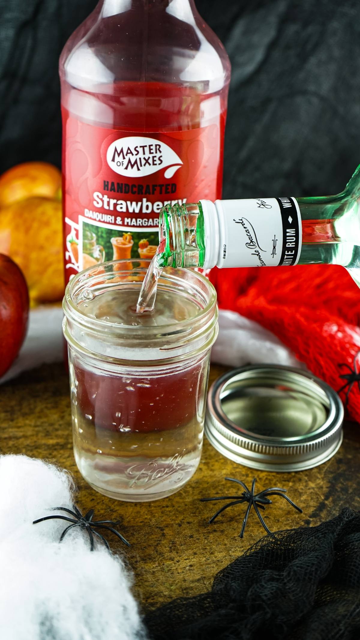 A bottle pours clear liquid into a jar beside strawberry mix, with Halloween decor like fake spiders—perfect for make ahead shots in a vial or syringe at your next adult Halloween party.