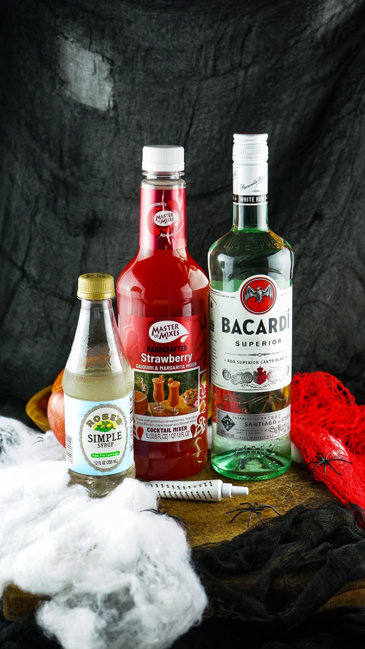 Three bottles—simple syrup, strawberry mixer, and Bacardi rum—await on a spooky Halloween setup, perfect for crafting Vampire Kiss Halloween Vial Shooter shots or make ahead shots in a syringe for your next adult party.