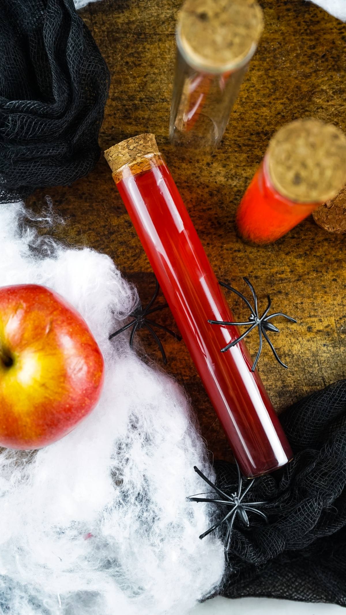 A red liquid in a corked test tube, perfect for Halloween rum shots, sits among plastic spiders, an apple, and fake cobwebs on a wooden surface—ideal for creative Halloween party ideas for adults.