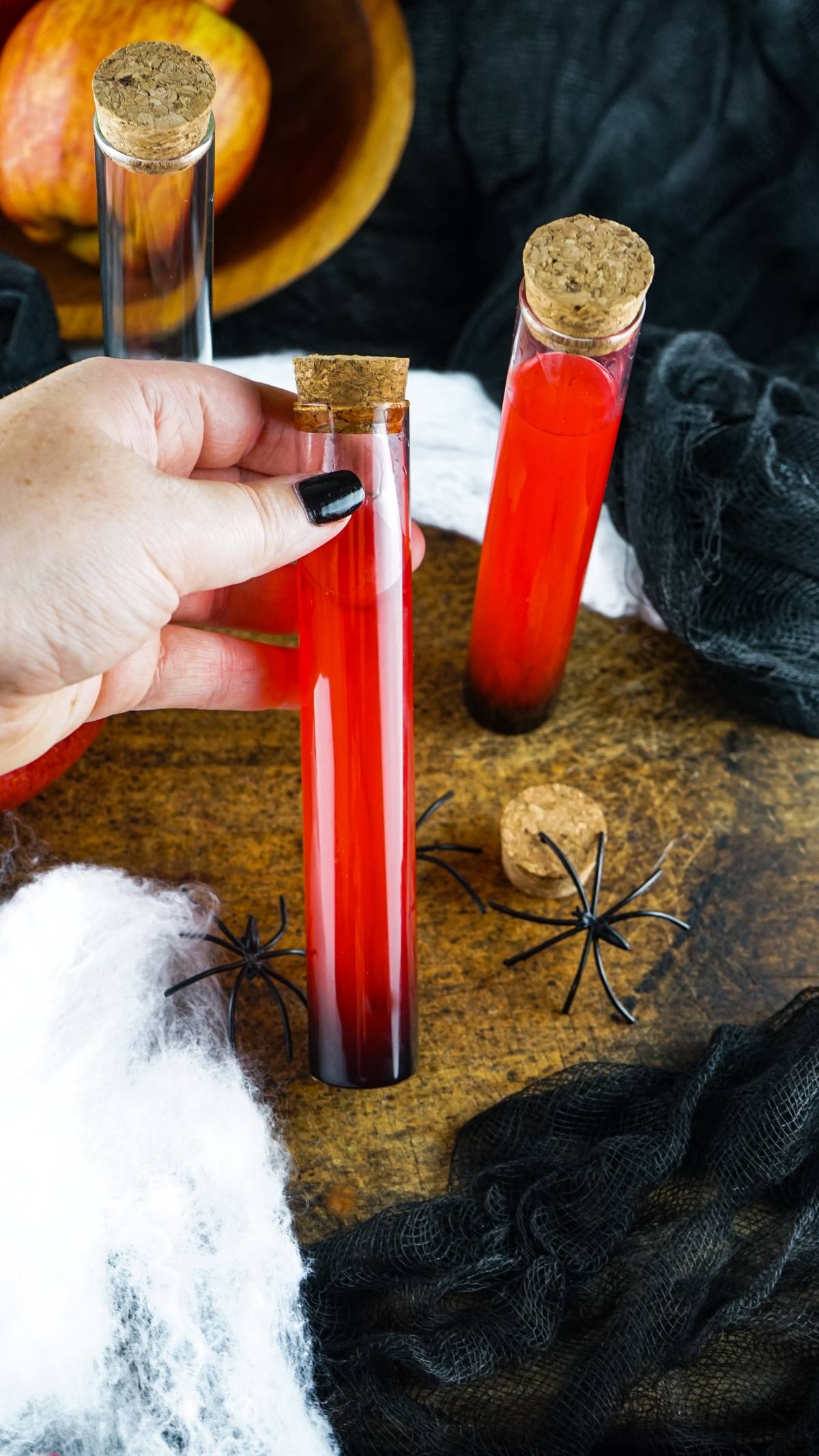 A hand holding a Vampire Kiss Halloween Vial Shooter—a red drink in a test tube with a cork—surrounded by spooky Halloween decorations. Perfect for make ahead shots in a vial or syringe!.