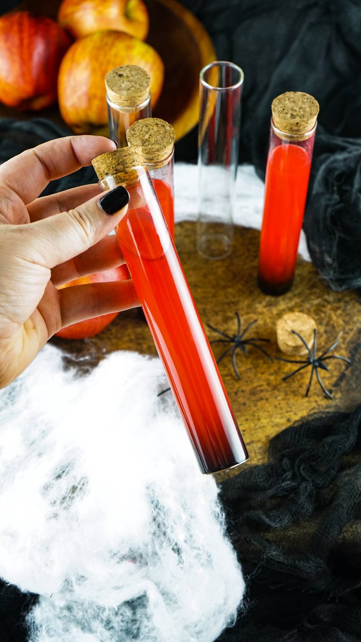 A hand holds a Vampire Kiss Halloween Vial Shooter filled with red liquid among test tubes, fake cobwebs, spiders, and apples—perfect inspiration for Halloween party ideas for adults.