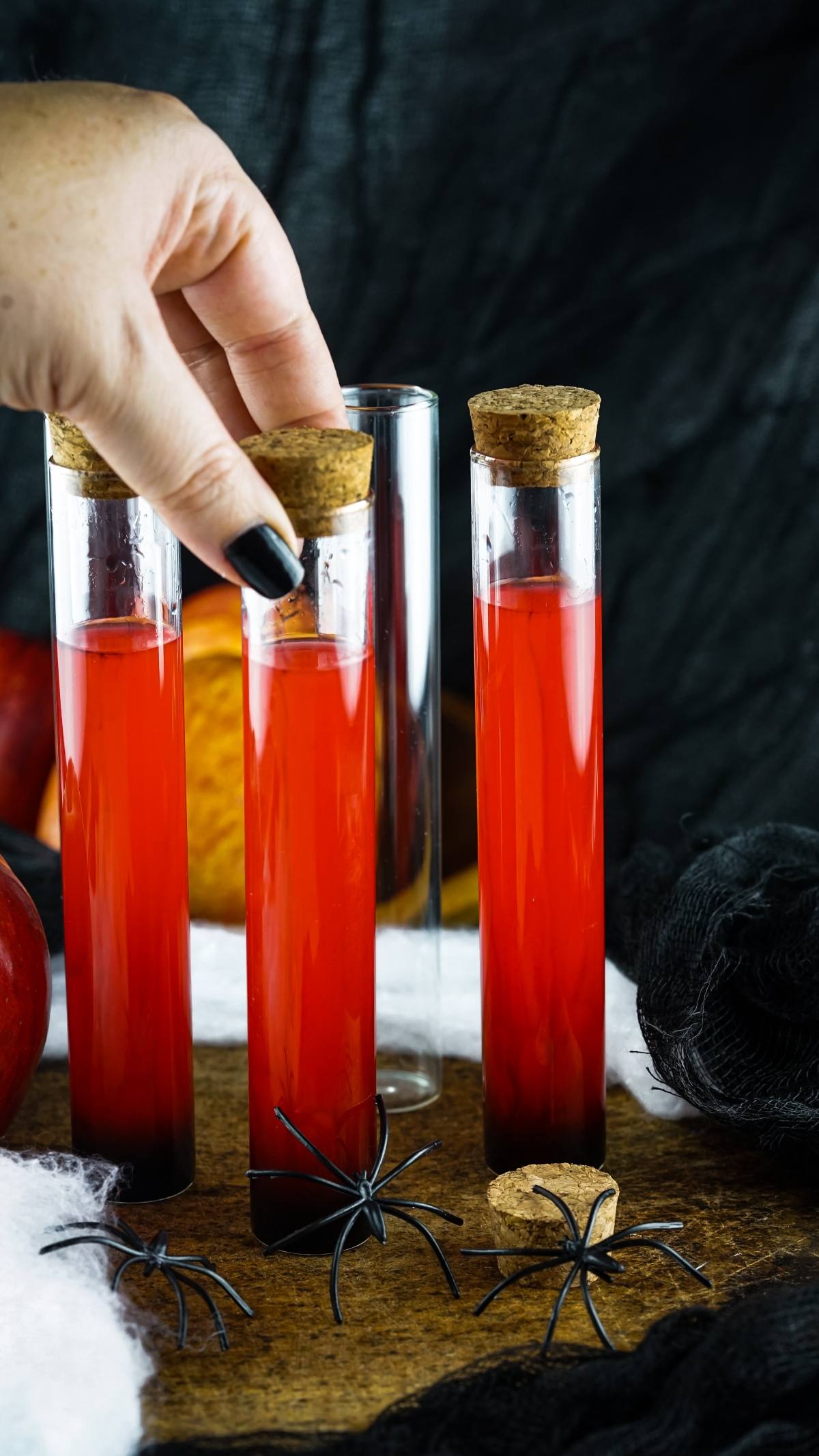 A hand places a cork in a test tube filled with red liquid, surrounded by fake spiders and Halloween decor—perfect inspiration for make ahead shots in a vial or syringe for your next Halloween party.