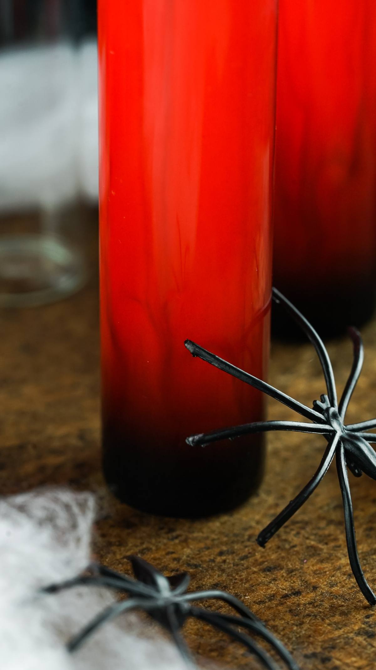 Tall glass of red and black layered drink with black plastic spiders on a brown surface—perfect for serving as Vampire Kiss Halloween Vial Shooter or make ahead shots in a vial or syringe.