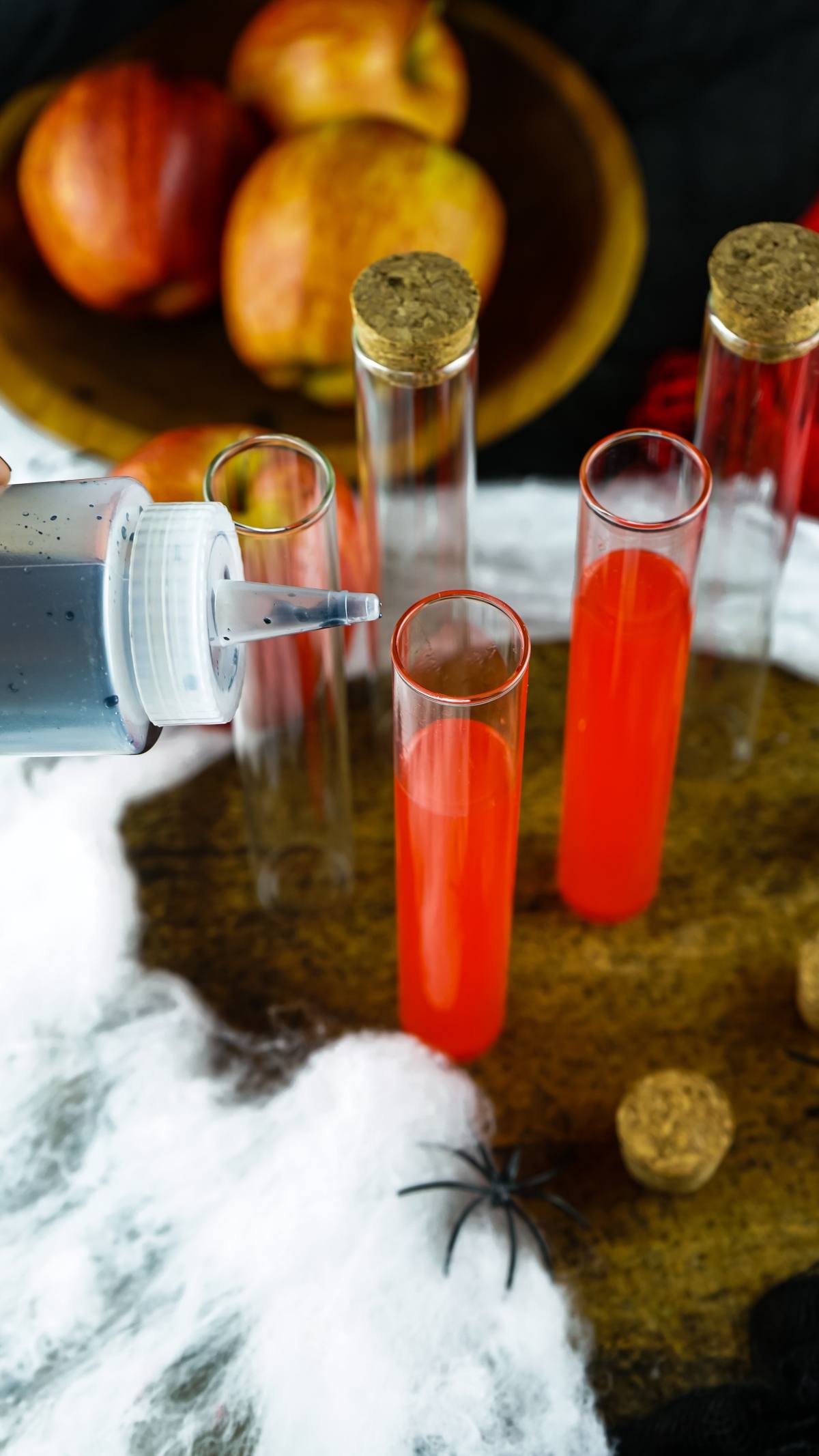 Three test tubes with red liquid, a dropper bottle, fake spider, apples, and white web decorations on a table—perfect make ahead shots in a vial or syringe for spooky Halloween party ideas for adults.
