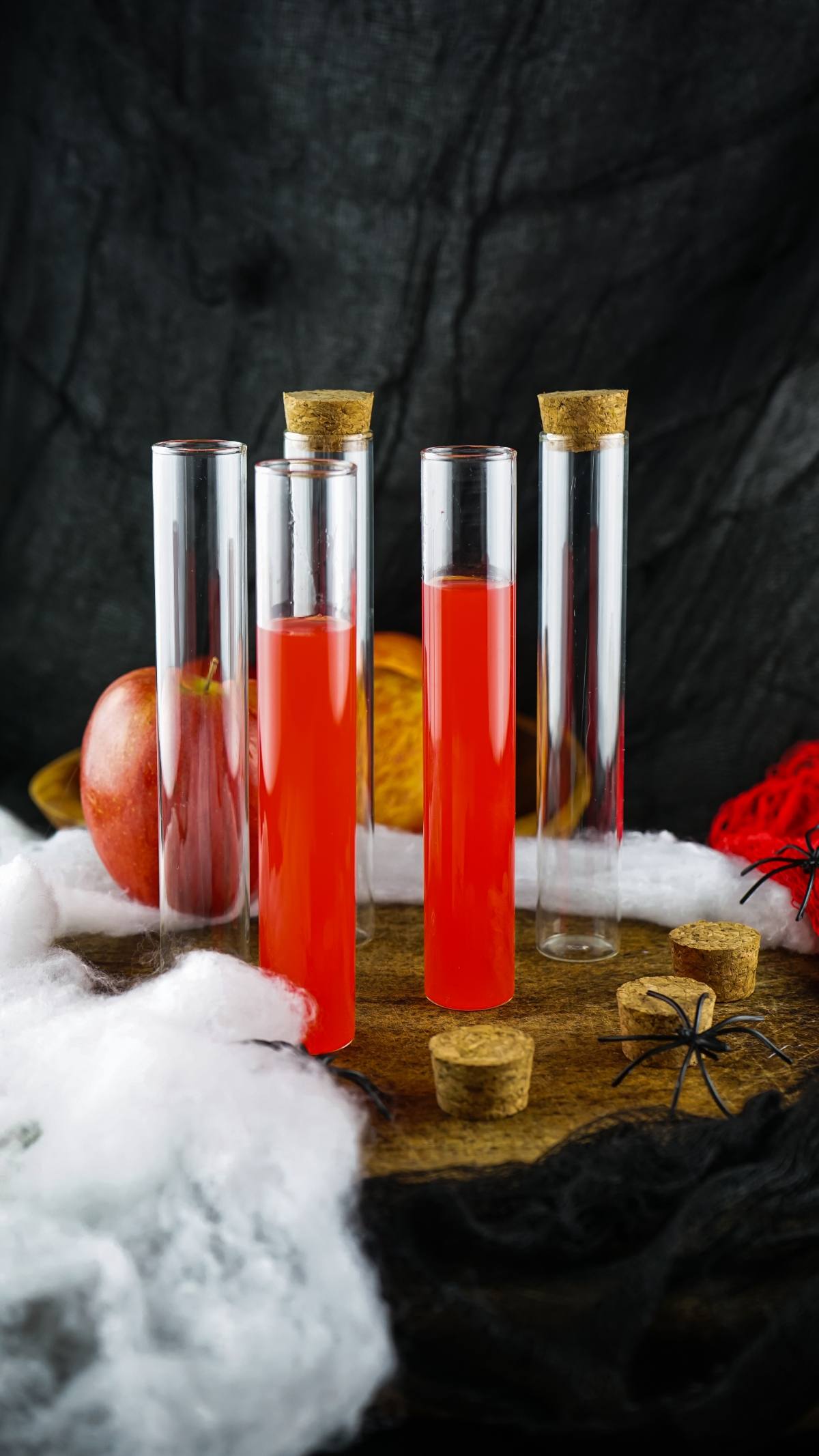 Three test tubes with red liquid and corks, surrounded by apples, a fake spider, and web on a wooden surface—perfect as Vampire Kiss Halloween Vial Shooters for unique alcoholic shots at your next adult Halloween party.