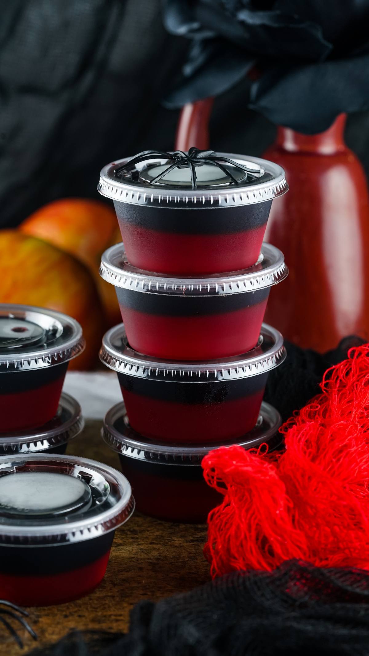 Four stacked red and black dessert cups with lids, perfect for Vampire Kiss Jello Shots, are surrounded by decorations and fruit—an eye-catching addition to your list of Halloween party ideas.