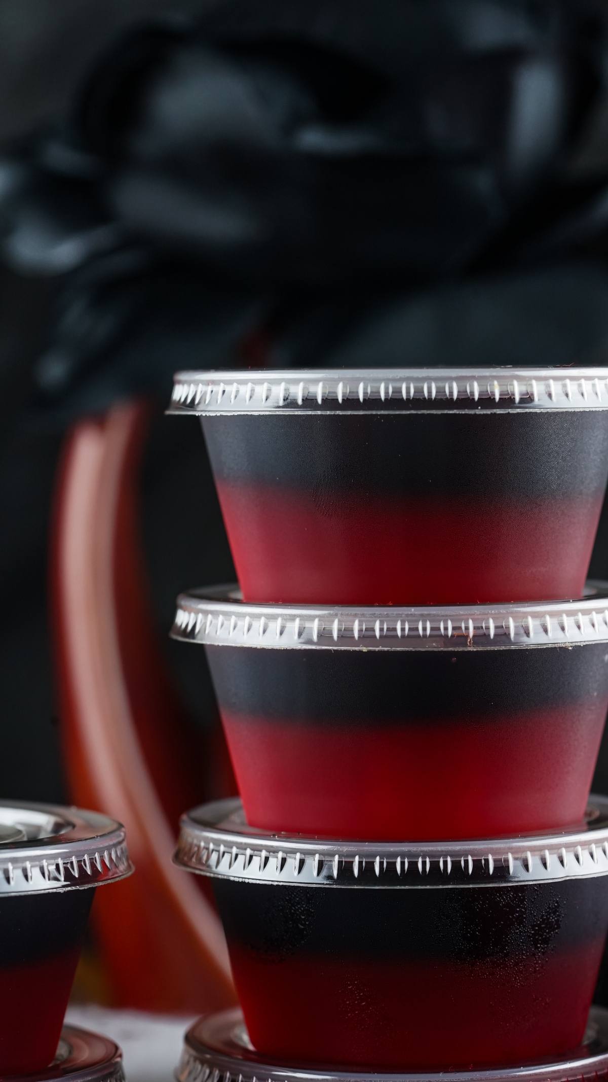 Stacked plastic cups filled with red and black layered gelatin, each with a silver lid, resemble a vampire blood jello shooter—perfect for those seeking how to make Halloween jello shots against a dark background.