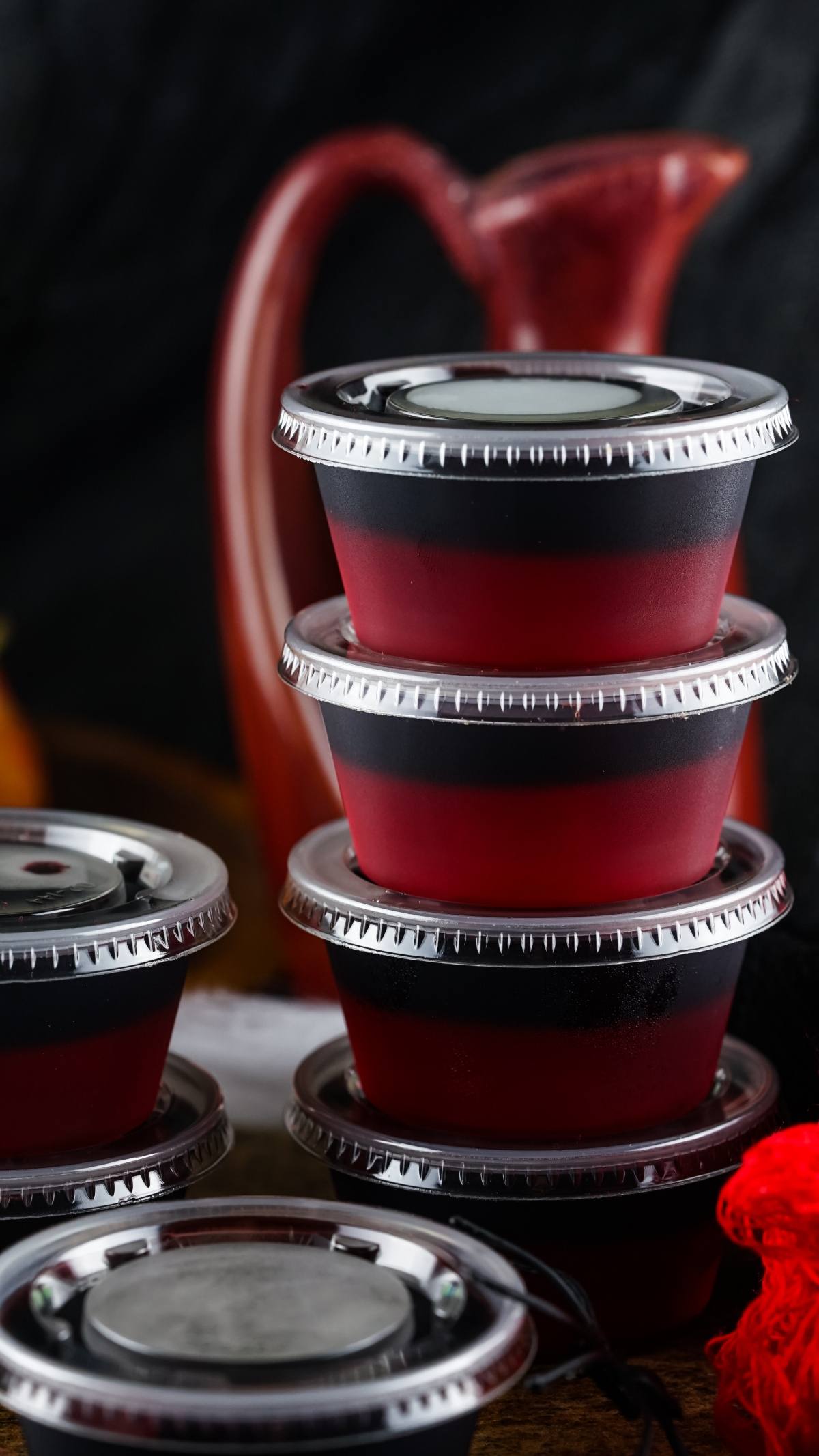 Four stacked red and black plastic cups with lids, perfect for serving Vampire Kiss Jello Shots or Halloween fireball shooters, with more cups and a red pitcher in the background.