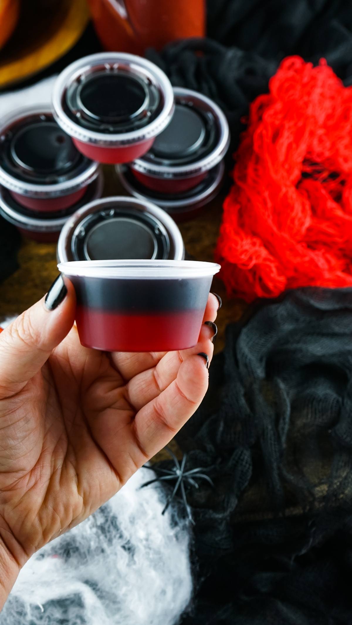 A hand holds a red and black Vampire Kiss Jello Shot in a plastic cup, with more cups and spooky Halloween decor in the background—perfect for unique Halloween party ideas.