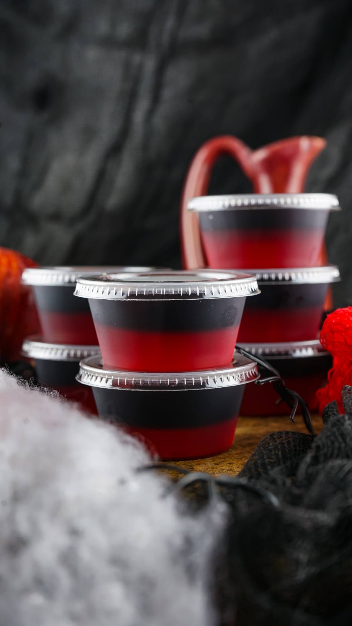 Stacked plastic cups of red and black layered gelatin, labeled as Vampire Kiss Jello Shots, with lids, set against a dark, festive background—perfect for creative Halloween party ideas.