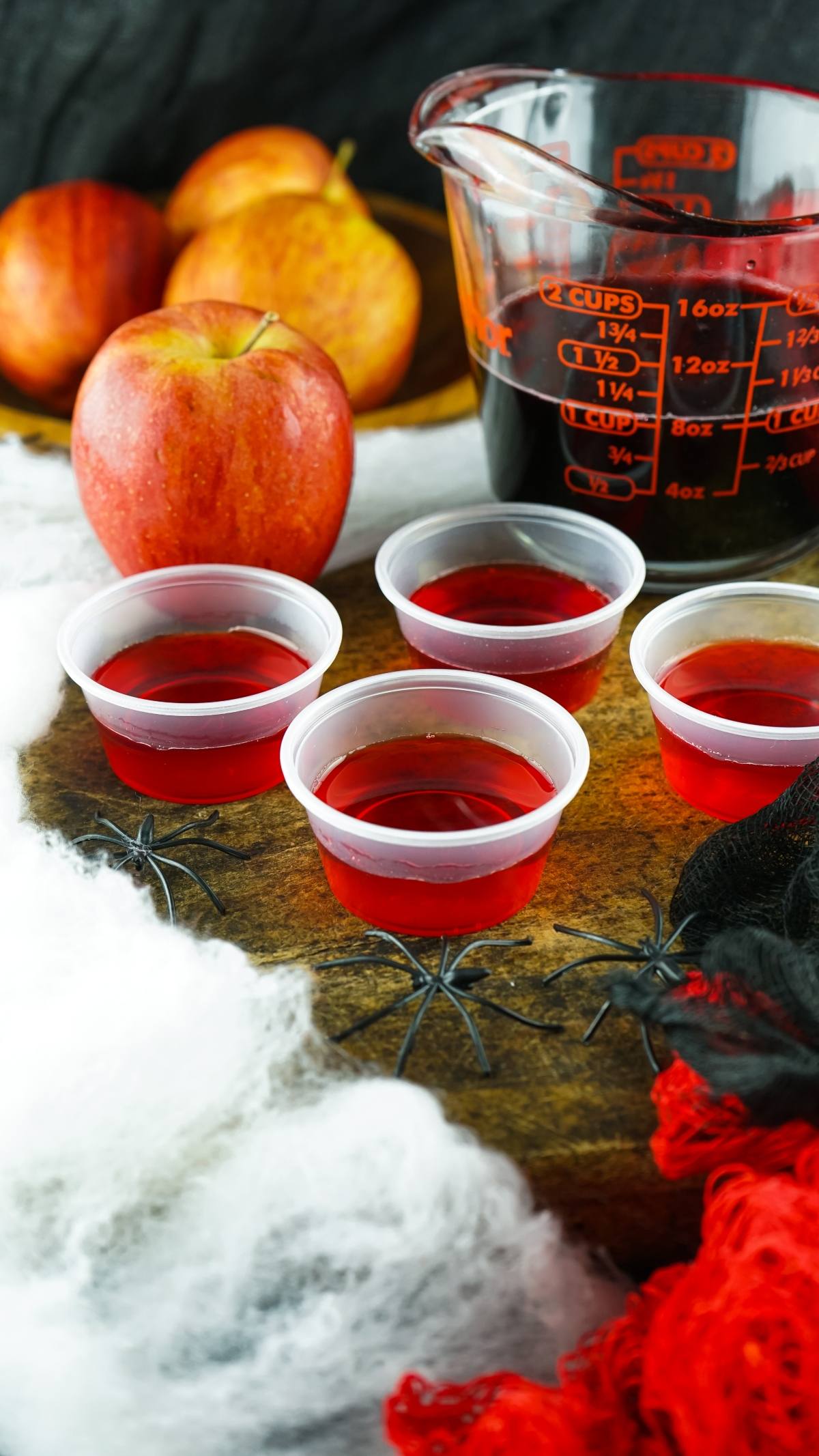 Four red gelatin shots in plastic cups sit on a table with apples, fake spider webs, and a measuring cup—a festive setup if you’re searching for how to make Halloween jello shots or fun Halloween party ideas.