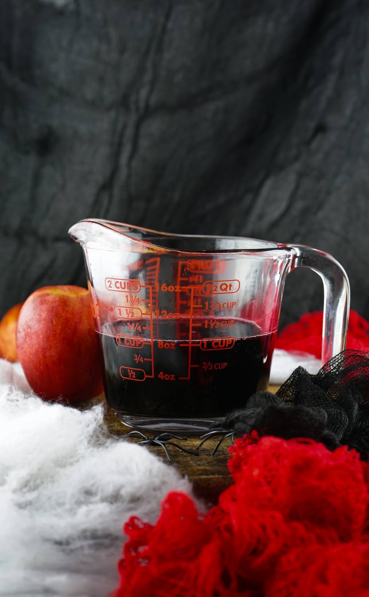 A measuring cup filled with dark red liquid, next to an apple, with red and black fabric in the background—perfect inspiration for Vampire Kiss Jello Shots or other creative Halloween party ideas.