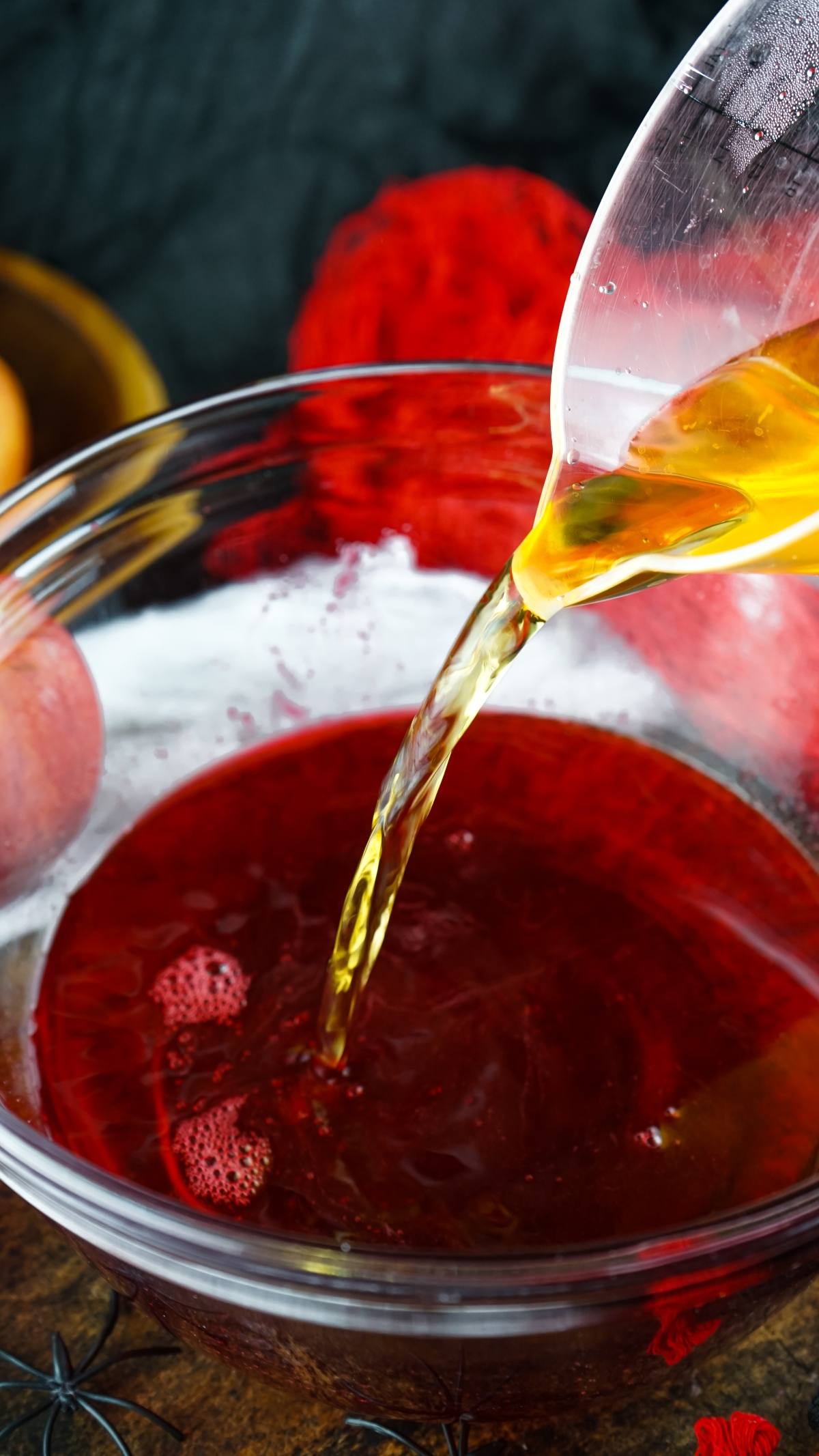 Golden liquid is poured from a measuring cup into a bowl of dark red vampire blood jello shooter, perfect for creative Halloween party ideas.