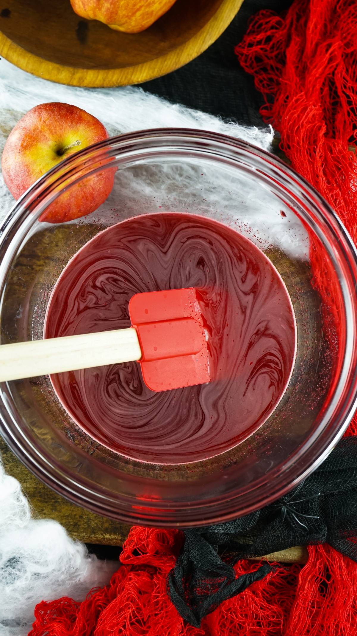 A glass bowl of dark red liquid being mixed with a red spatula, next to an apple and red cloth—perfect for prepping a vampire blood jello shooter or learning how to make Halloween jello shots.