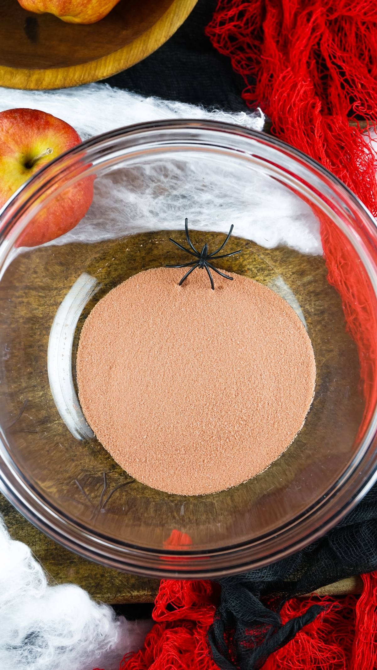A glass bowl with orange powder, perfect for serving Vampire Kiss Jello Shots, is topped with a black spider decoration and surrounded by apples and red netting.