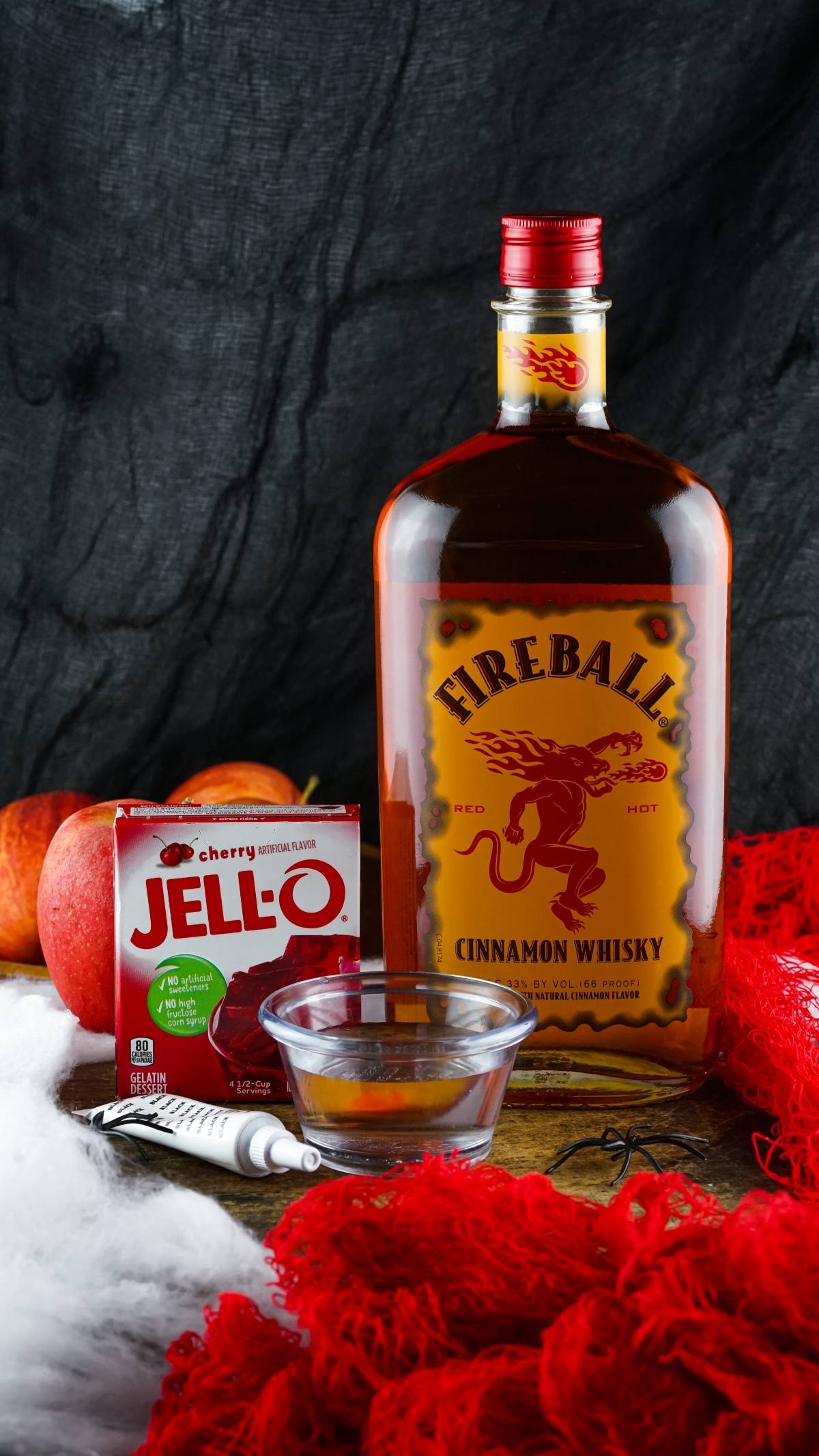 Fireball whisky, cherry Jell-O, apples, and a syringe in a glass bowl on a wooden table make the perfect vampire blood jello shooter—one of the best Halloween party ideas.