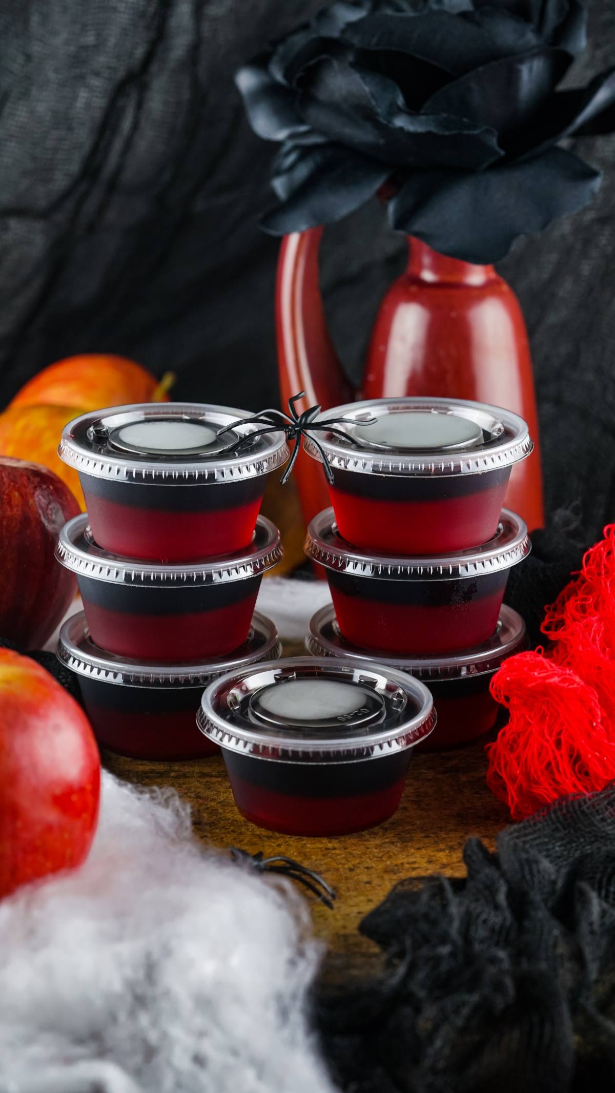 Six small red containers of Vampire Kiss Jello Shots are stacked on a table with apples, a red vase, and black decorative flowers nearby—perfect for creative Halloween party ideas.