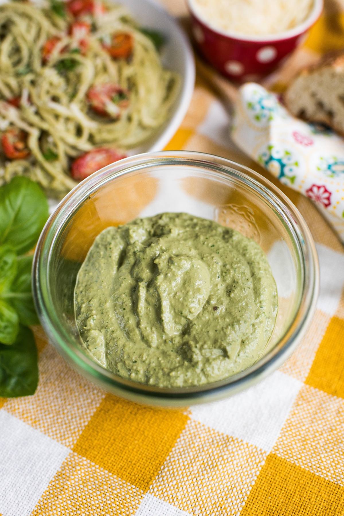 A glass bowl of vegan pesto sauce sits on a yellow and white checkered tablecloth.