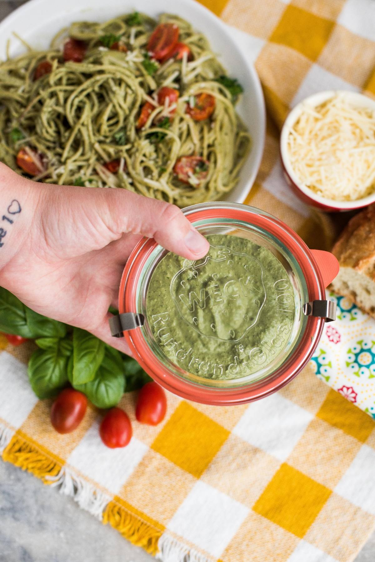 A hand holds a jar of vegan pesto above a table with pasta, cheese, bread, basil, and tomatoes.