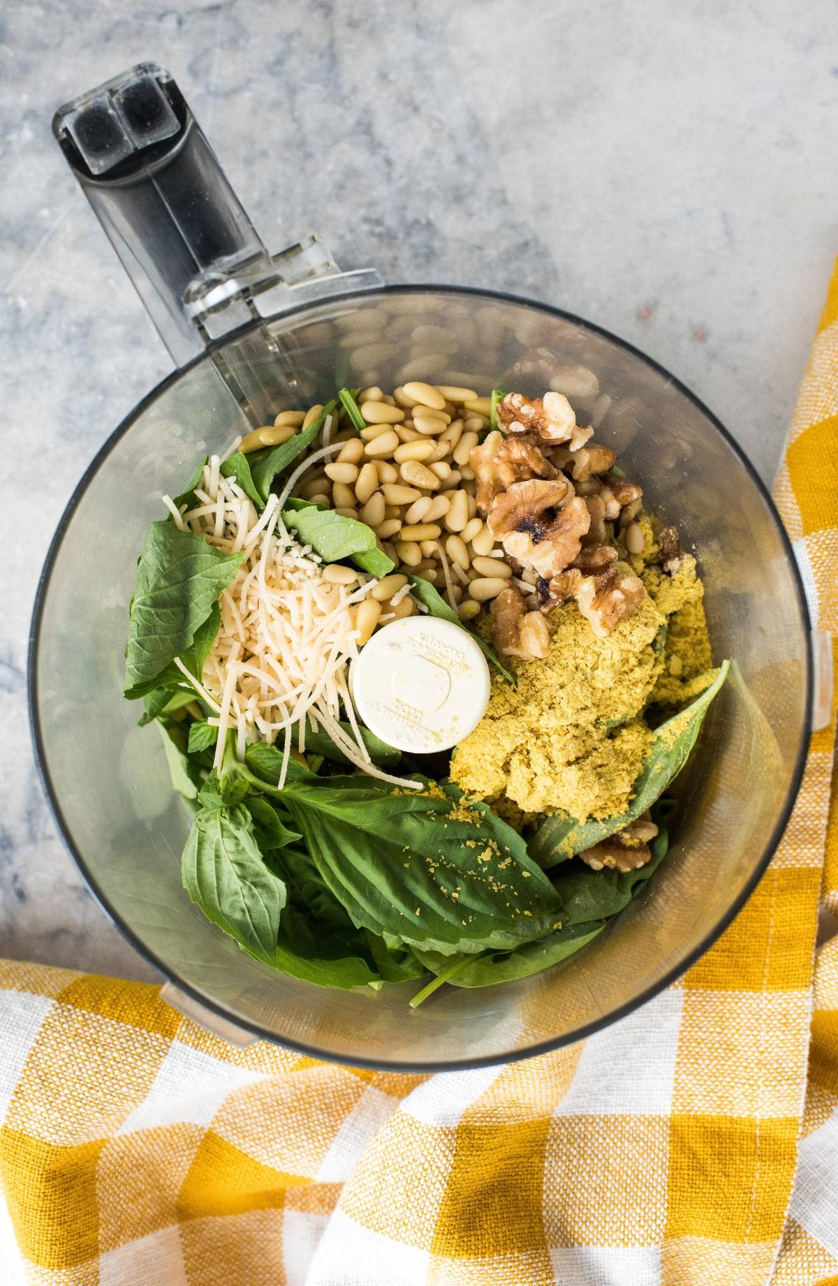 Food processor bowl with basil, pine nuts, walnuts, Parmesan, and nutritional yeast, ready to make vegan pesto sauce.