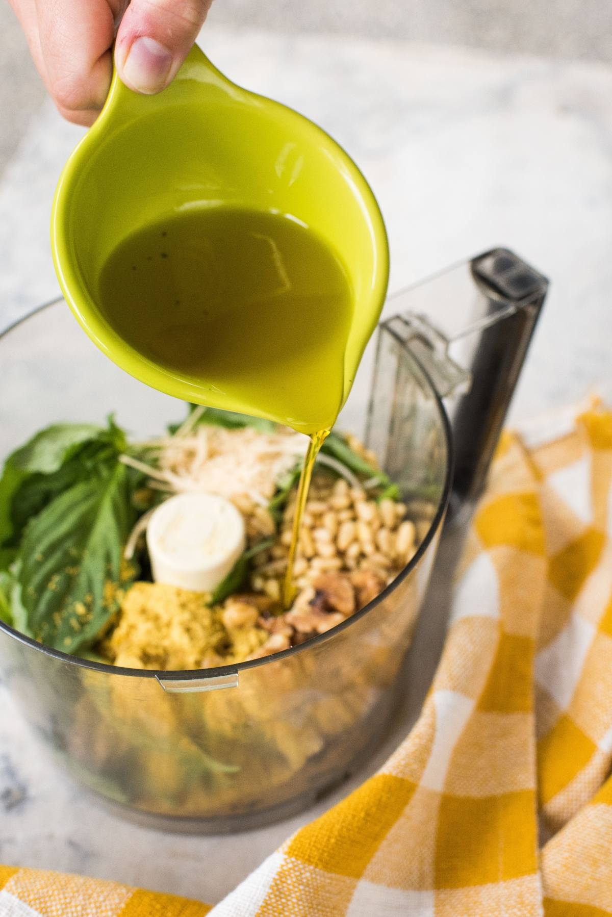 Olive oil is being poured into a food processor with basil, nuts, and cheese to create a delicious vegan pesto sauce, next to a yellow checkered cloth.