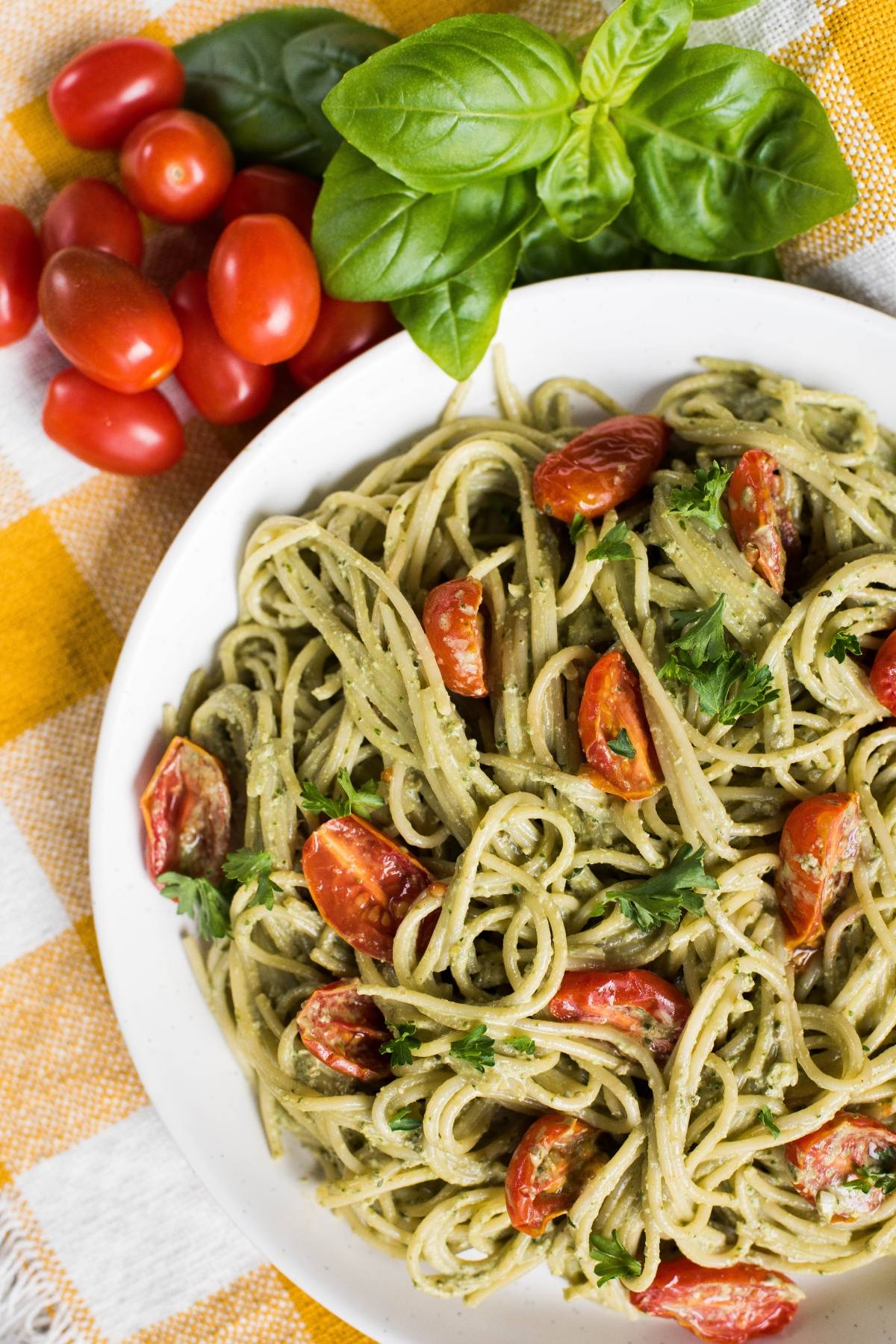 Plate of spaghetti with vegan pesto sauce and cherry tomatoes, garnished with parsley, next to fresh basil and tomatoes.