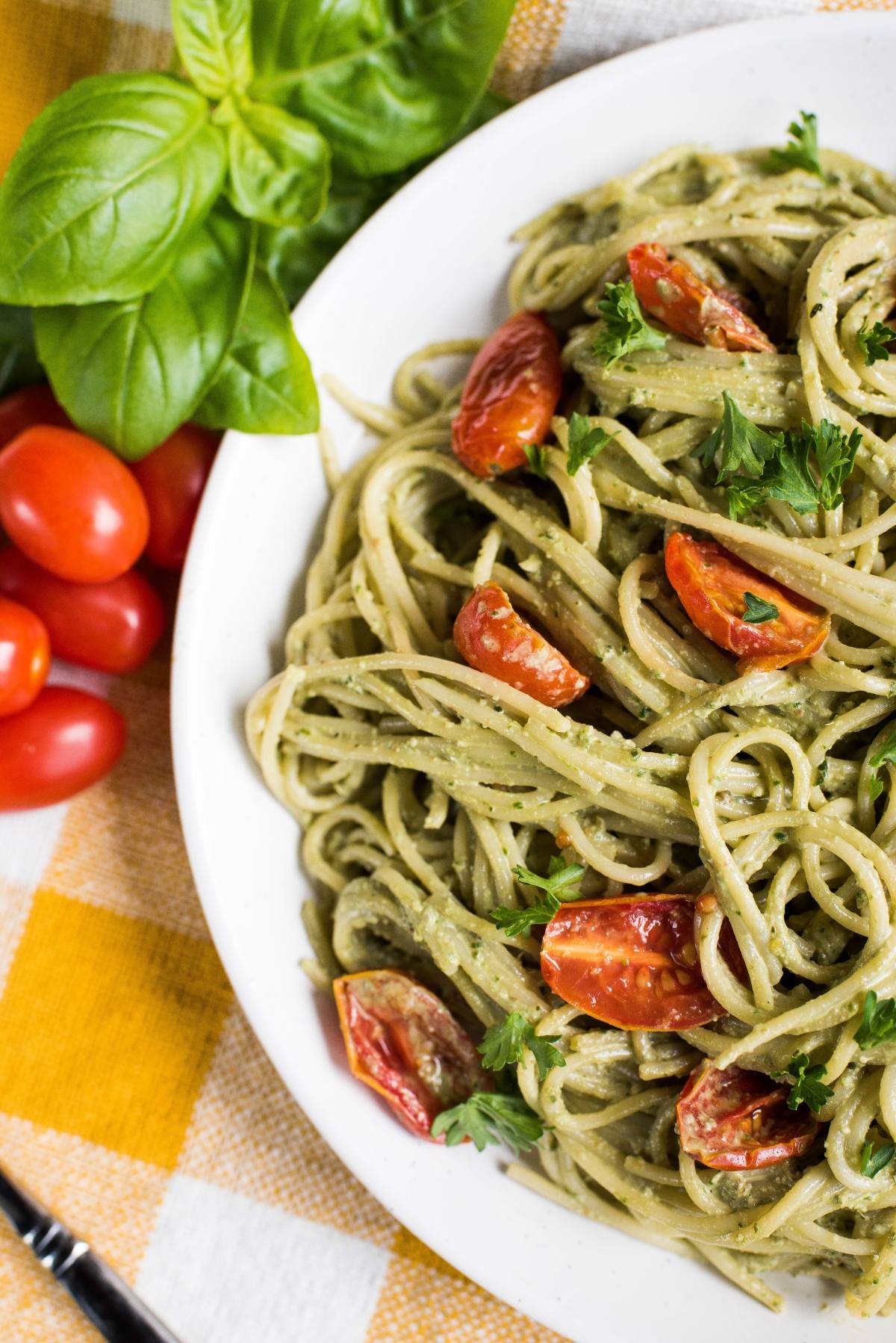 A plate of pasta tossed in vegan pesto sauce with cherry tomatoes, garnished with parsley and fresh basil, on a checkered tablecloth.