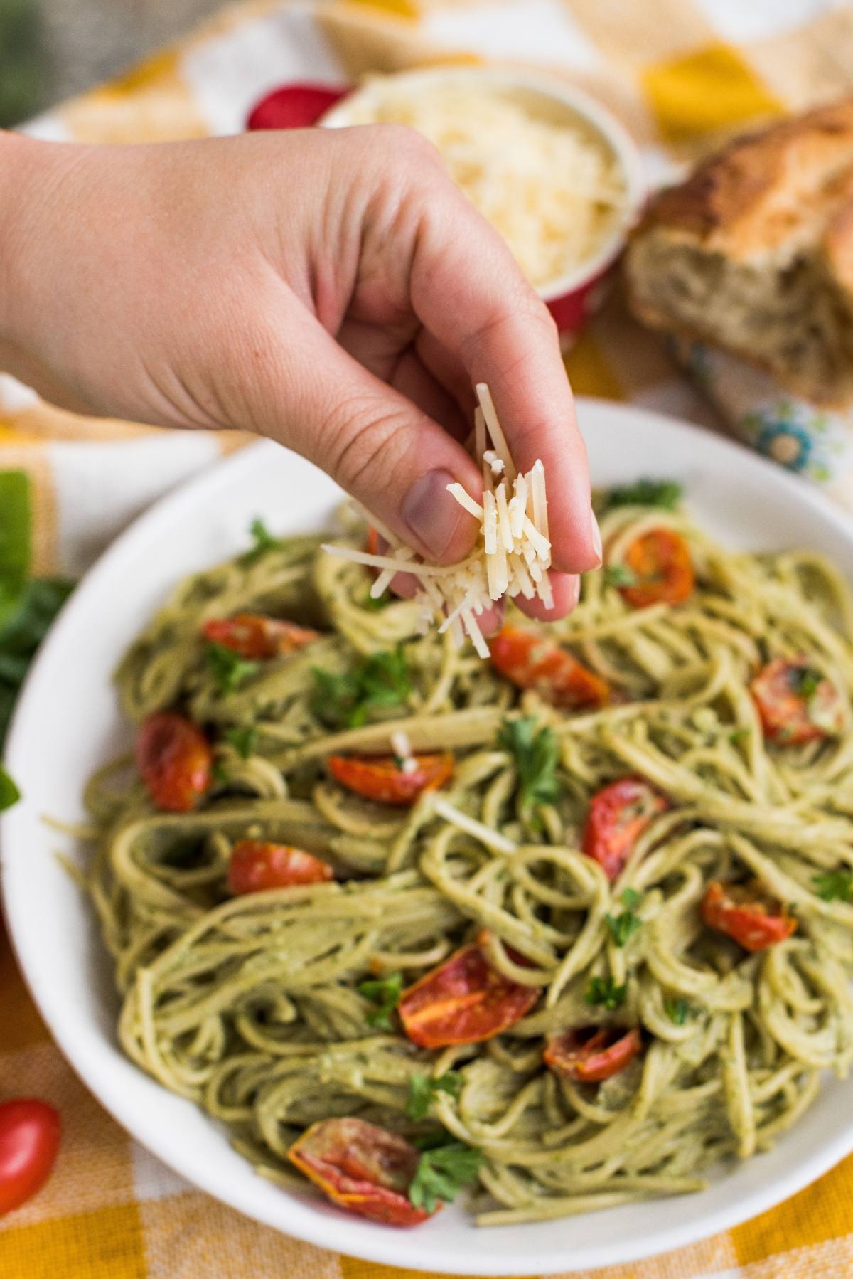A hand sprinkles shredded cheese over a plate of pasta with vegan pesto sauce and cherry tomatoes.