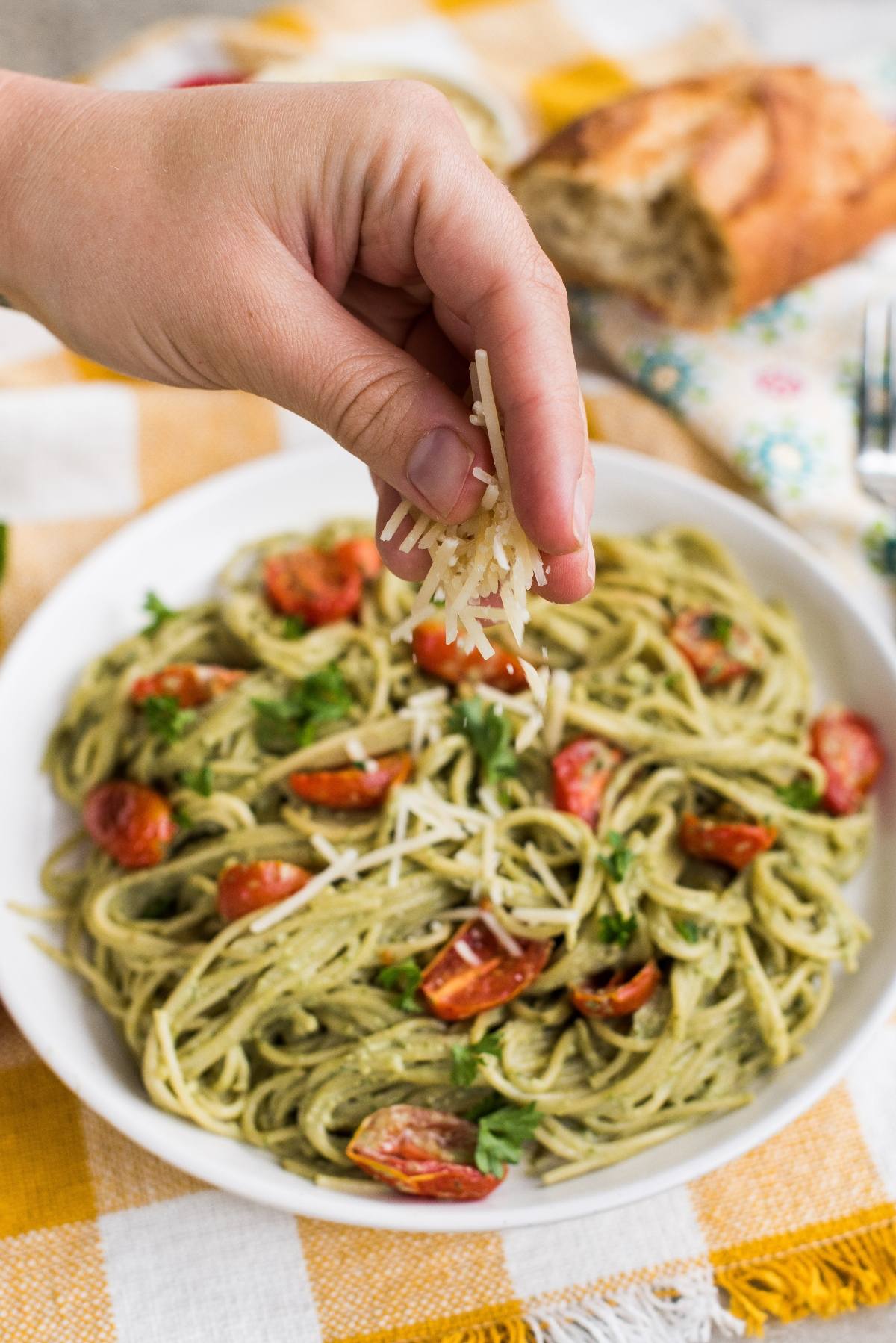 A hand sprinkles grated cheese over a plate of pasta tossed in vibrant vegan pesto sauce, garnished with cherry tomatoes and herbs.