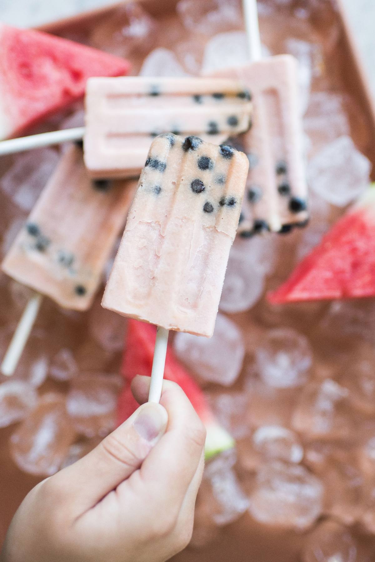 Hand holding a popsicle with chocolate chips, with more watermelon boba popsicles, ice, and watermelon slices in the background.